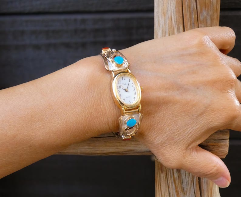 May include: Gold-toned wristwatch with an oval face and a white dial. The watch band features silver and turquoise accents. The watch is worn on a wrist, showcasing the design and craftsmanship of the timepiece.