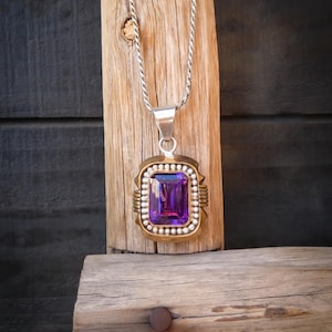 May include: A silver necklace with a rectangular purple gemstone pendant. The pendant is set in a gold-toned frame with a beaded border. The necklace is displayed on a wooden surface.