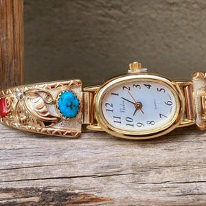 May include: Gold-toned watch with an oval face and a white dial featuring black numerals. The watch band is adorned with turquoise and red stones, and intricate gold filigree designs. The watch is displayed on a weathered wooden surface.