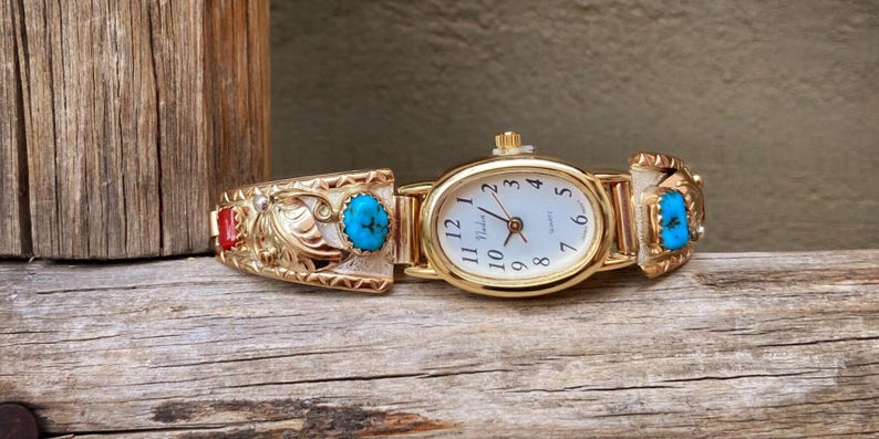 May include: Gold-toned watch with an oval face and a white dial featuring black numerals. The watch band is adorned with turquoise and red stones, and intricate gold filigree designs. The watch is displayed on a weathered wooden surface.