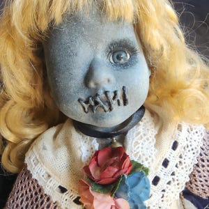 Op de afbeelding: Een vintage pop met een grijs gezicht, een gestikte mond en één zichtbaar blauw oog. De pop heeft blond krullend haar en draagt een crèmekleurige jurk met een bloemenaccent. Een zwarte choker zit om de nek van de pop.