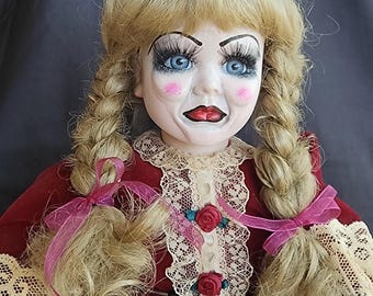 May include: A vintage doll with a porcelain face, blue eyes, and long eyelashes. The doll has blonde hair styled in braids with pink ribbons. It wears a red velvet dress with lace trim and a cream-colored satin bow.
