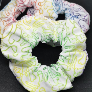 May include: White scrunchie with a rainbow pattern of various shapes and lines. The scrunchie is made of fabric and has a gathered look.