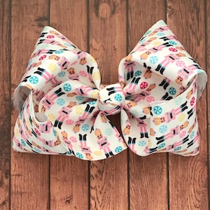 May include: A large white bow with a pink and white pattern of gingerbread men and candy canes. The bow is tied with a knot in the center and has a white background. The text "Stephanie's Bows" is written in white cursive on a brown wooden background.