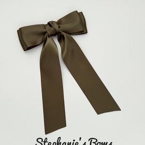 May include: Olive green satin bow with long, flowing tails. The bow is tied in the center and has a classic shape. The text "Stephanie's Bows" is written in a cursive font at the bottom.