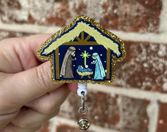 May include: A gold glitter Christmas nativity scene badge reel with a blue background and a white star. The scene depicts Mary, Joseph, and baby Jesus in a manger.