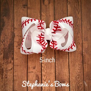 May include: A white and red baseball-themed hair bow with red stitching detail. The bow is 5 inches wide.