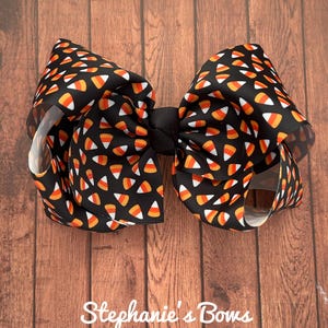 May include: A large, decorative hair bow featuring a black background with a repeating pattern of candy corn in orange, yellow, and white. The bow is tied with a black center knot and has a layered design. The text "Stephanie's Bows" is at the bottom.