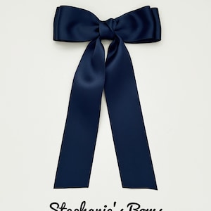 May include: A dark navy blue satin hair bow featuring a central knot and two long, smooth ribbon tails hanging downwards. The text "Stephanie's Bows" is printed in a black cursive script font at the bottom of the image below the ribbon tails.