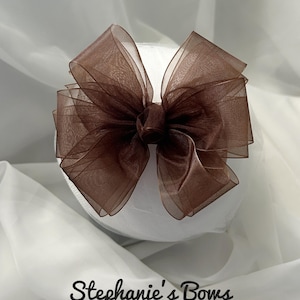 May include: A large, brown, sheer bow with multiple layers, centered on a white, rounded form. The bow is made of a delicate, possibly organza-like material. The text "Stephanie's Bows" is written in a cursive font at the bottom.