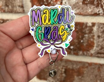 May include: A white Mardi Gras themed badge holder with a purple, green, and gold glitter design. The badge holder has a clear retractable cord.