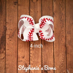 May include: A white 4-inch hair bow with a red baseball stitch design. The bow is on a wood background. "Stephanie's Bows" is written below the bow.