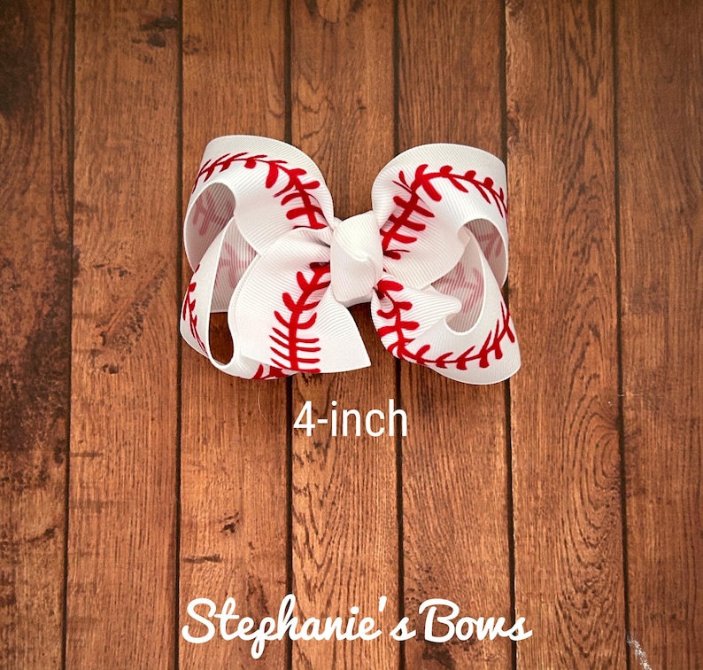 May include: A white 4-inch hair bow with a red baseball stitch design. The bow is on a wood background. "Stephanie's Bows" is written below the bow.