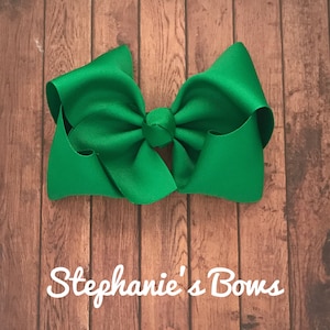 May include: A large green bow made from grosgrain ribbon. The bow is tied with a knot in the center and has a slightly ruffled texture. The bow is photographed against a woodgrain background.