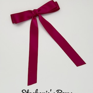 May include: A magenta satin bow with long, flowing tails. The bow is tied in the center and lies on a white background. The text "Stephanie's Bows" is written in cursive below the bow.