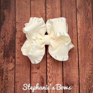 May include: Ivory-colored hair bow with ruffled edges, centered with a matching ribbon. The bow is set against a dark wood grain background. The text "Stephanie's Bows" is visible at the bottom of the image.