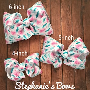 May include: Three white hair bows with a Christmas tree pattern in pink and teal. The bows are labeled 4-inch, 5-inch, and 6-inch. The bows are on a wooden surface. The text "Stephanie's Bows" is at the bottom.