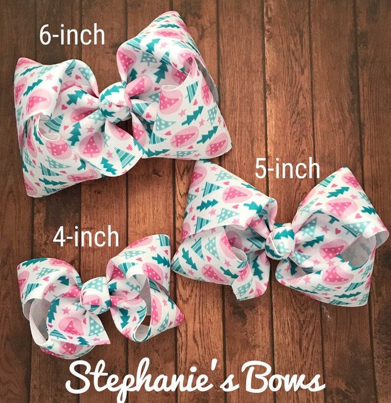 May include: Three white hair bows with a Christmas tree pattern in pink and teal. The bows are labeled 4-inch, 5-inch, and 6-inch. The bows are on a wooden surface. The text "Stephanie's Bows" is at the bottom.