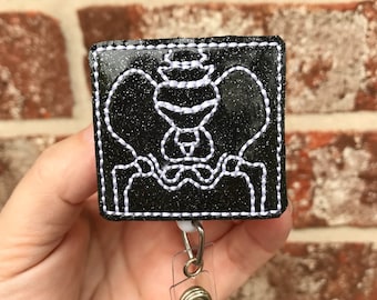 May include: Black glitter badge holder with a white embroidered image of a human pelvis. The badge holder is attached to a clear retractable badge reel with a silver clip.