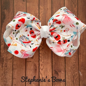 May include: A white bow with a Nutcracker themed print. The bow features a white center and is decorated with images of Nutcracker soldiers, ballerinas, and other holiday characters. The bow is on a wooden background.
