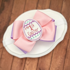 May include: A pink and purple bow headband with an embroidered white egg that says "My 1st Easter" in pink and purple lettering.