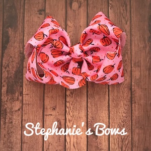 May include: A large pink hair bow with a pumpkin print. The bow is made of grosgrain ribbon and has a wired edge. The bow is tied in a classic bow shape and has a loop at the top for attaching to hair.