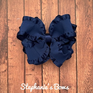 May include: A large, navy blue hair bow with ruffled edges. The bow is centered on a wooden background. The text "Stephanie's Bows" is written in cursive at the bottom of the image.