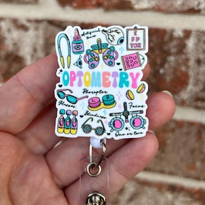 May include: A white and pink retractable badge holder with a colorful illustration of various optometry-related items, including glasses, an eye chart, and the word "OPTOMETRY".