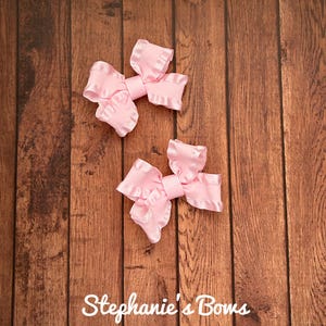 May include: Two light pink hair bows with ruffled edges and a center band are displayed on a wooden surface. The bows are made of a satin-like material. The text "Stephanie's Bows" is at the bottom of the image.