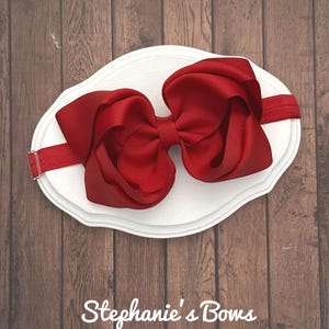 May include: A large, vibrant red bow headband. The bow is made of layered fabric and is attached to a matching red elastic band. The headband is displayed on a white, scalloped-edge surface. The text "Stephanie's Bows" is visible.