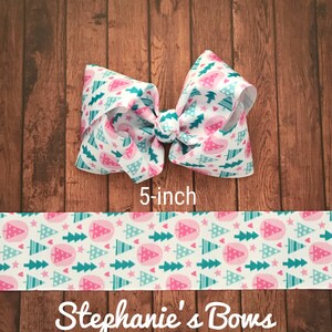 May include: A 5-inch hair bow with a white background and a repeating pattern of pink and teal Christmas trees and stars. The bow is tied in the center. The text "Stephanie's Bows" is at the bottom.