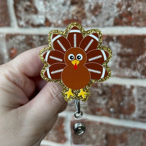May include: A gold glitter turkey with footballs for feathers. The turkey has a red wattle and a brown body. The turkey is attached to a clear plastic retractable badge holder with a silver clip.