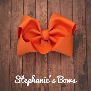 May include: Bright orange hair bow made of grosgrain ribbon. The bow is centered and has a knotted center. The background is a wooden surface. The text "Stephanie's Bows" is written in white script at the bottom.