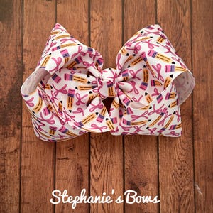 May include: A large, white hair bow with a repeating pattern of yellow pencils and pink ribbons. The bow is tied in the center and has a layered design. The text "Stephanie's Bows" is at the bottom.