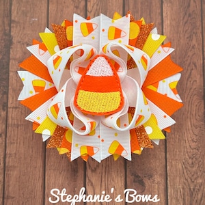 May include: A handmade Halloween hair bow featuring a candy corn design. The bow is made of white, orange, and yellow ribbons, with a candy corn applique in the center. The bow has a glittery orange accent and the text "Stephanie's Bows".