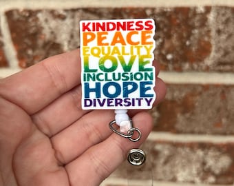 Könnte beinhalten: Ein rechteckiges Ausweis-Rollband mit den Worten "KINDNESS, PEACE, EQUALITY, LOVE, INCLUSION, HOPE, DIVERSITY" in einem Regenbogenverlauf. Das Rollband hat ein weißes Kunststoffgehäuse, einen Metallclip und einen transparenten Kunststoffriemen.