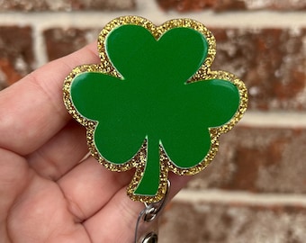 May include: A green shamrock badge reel with a gold glitter border. The badge reel has a retractable cord and a metal clip. The shamrock is a symbol of good luck and is often associated with St. Patrick's Day.