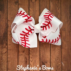 May include: White hair bow with a red baseball stitch design.  "Stephanie's Bows" is written in the bottom of the image.