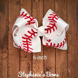 May include: A white and red baseball-themed hair bow with red stitching. The bow is 6 inches in diameter.  "Stephanie's Bows" is written below the bow.