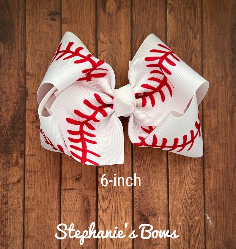 May include: A white and red baseball-themed hair bow with red stitching. The bow is 6 inches in diameter.  "Stephanie's Bows" is written below the bow.