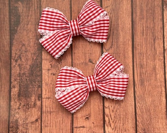 Könnte beinhalten: Zwei rot-weiß karierte Haarschleifen mit weißer Spitzenborte auf einem Holzgrund. Die Schleifen sind in klassischer Schleifenform gebunden. Der Text "Stephanie's Bows" befindet sich unten.
