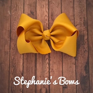 May include: A large, mustard-yellow hair bow made of grosgrain ribbon. The bow is centered and knotted, with large loops on either side. The background is a wooden surface. The text "Stephanie's Bows" is at the bottom.