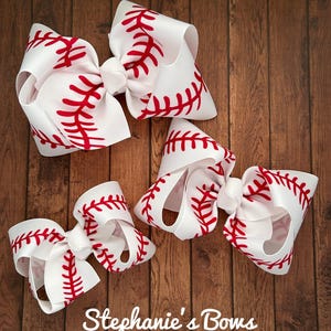 May include: Three white hair bows with red baseball stitching. The bows are arranged in a triangular shape on a wooden background. The largest bow is at the top, and the smallest bow is at the bottom. The text "Stephanie's Bows" is at the bottom of the image.
