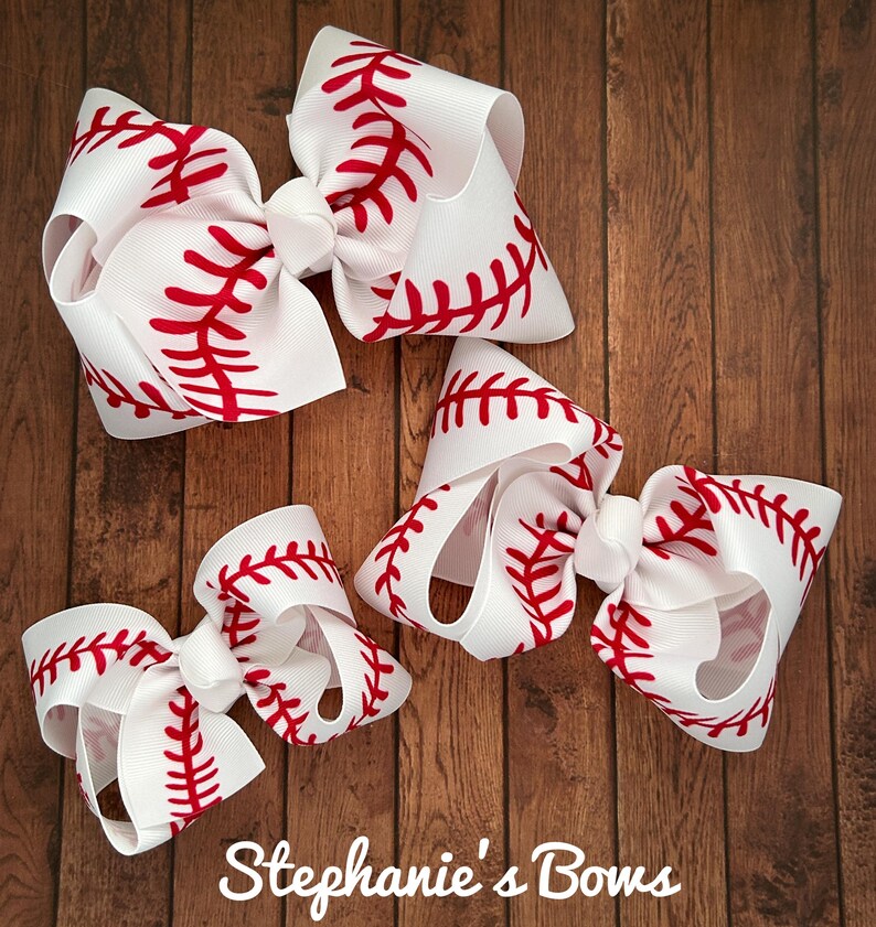 May include: Three white hair bows with red baseball stitching. The bows are arranged in a triangular shape on a wooden background. The largest bow is at the top, and the smallest bow is at the bottom. The text "Stephanie's Bows" is at the bottom of the image.