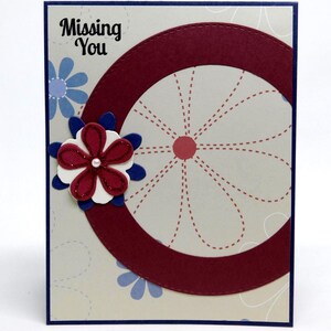 May include: A greeting card with a navy blue border and a cream-colored background. The card features a large burgundy circle and a flower with burgundy, white, and blue petals. The words "Missing You" are at the top.