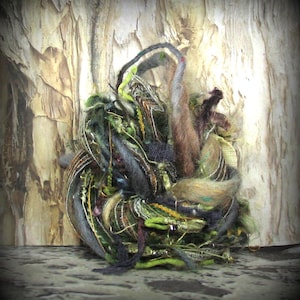 May include: A circular fiber art piece composed of tangled yarn, thread, and fabric strips in shades of forest green, olive, charcoal gray, and brown. The textured bundle features metallic gold accents, fuzzy wool fibers, and various woven strands.