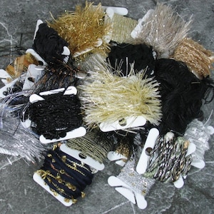 May include: A collection of assorted decorative ribbons and novelty yarns wound onto white plastic bobbins. The assortment includes metallic gold, silver, and black textures, featuring fringed, sequined, and twisted cord styles in various lengths.