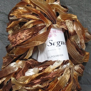 May include: A skein of ribbon yarn in shades of metallic bronze, copper, and cream. The yarn features a crinkled, shimmering texture. A white paper label is wrapped around the center, printed with the text Handsetter Yarns Segue and 100% Polyamide.