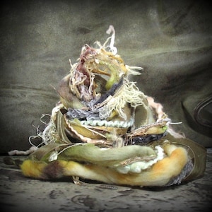 May include: A pile of yarn, fabric scraps, and beads in shades of brown, beige, and green. The materials are arranged in a loose, organic shape, resembling a nest or a small sculpture.