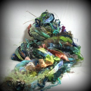 May include: A pile of colorful craft yarn in shades of green, blue, and brown. The yarn includes ribbons, metallic threads, and textured fibers. The arrangement is dense and layered, with a variety of textures and colors.